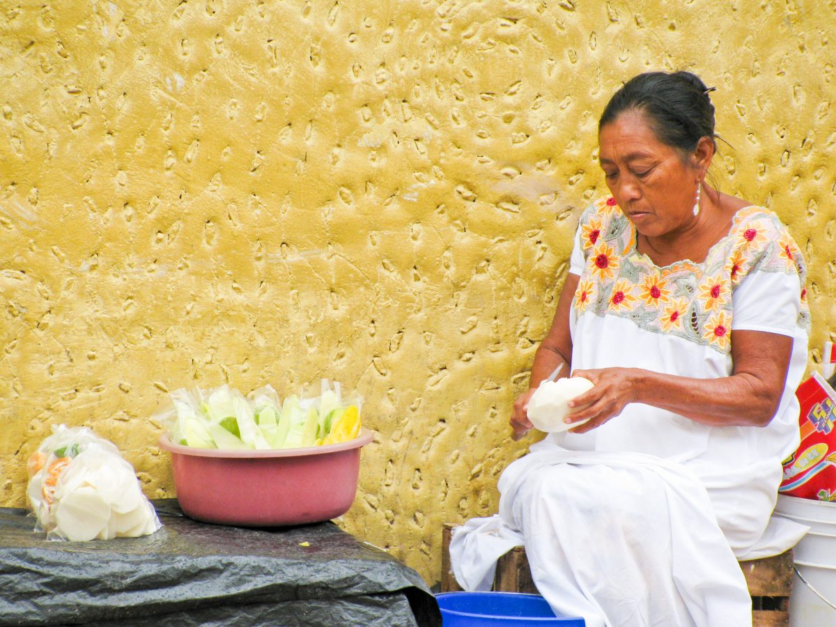 Mayan woman in white peeling mangos in front of yellow wall