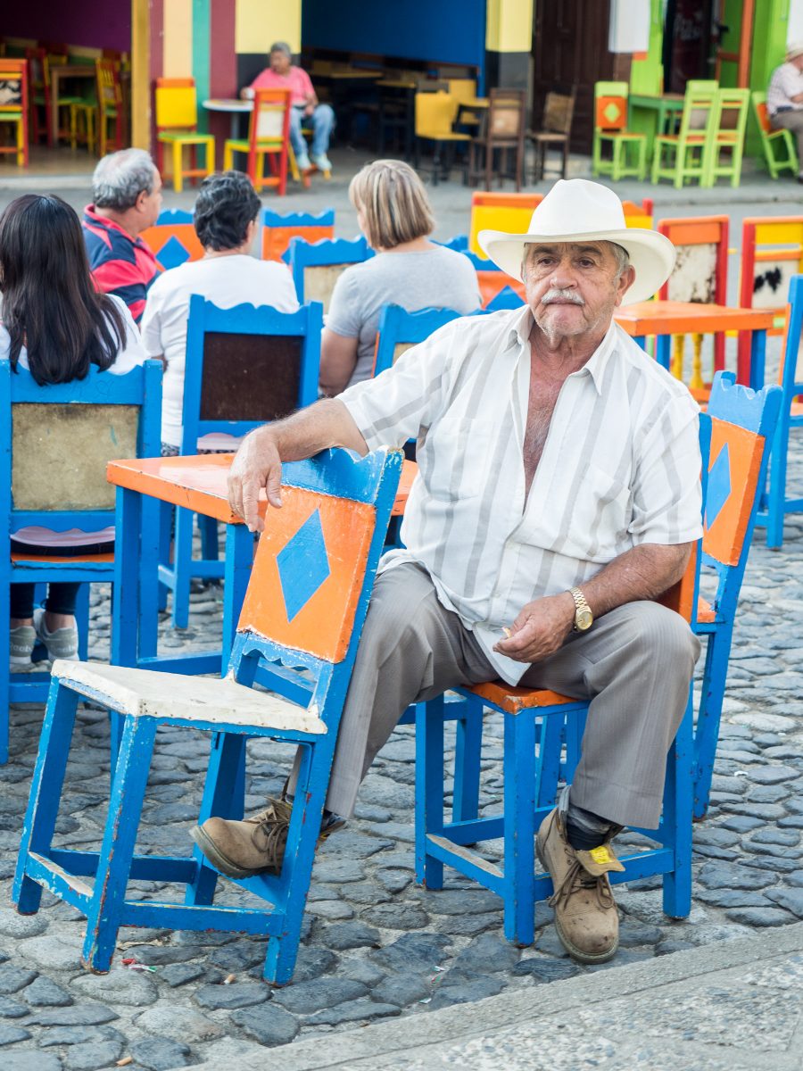 Older man in cowboy hat lounges on blue and orange cafe stools in plaza