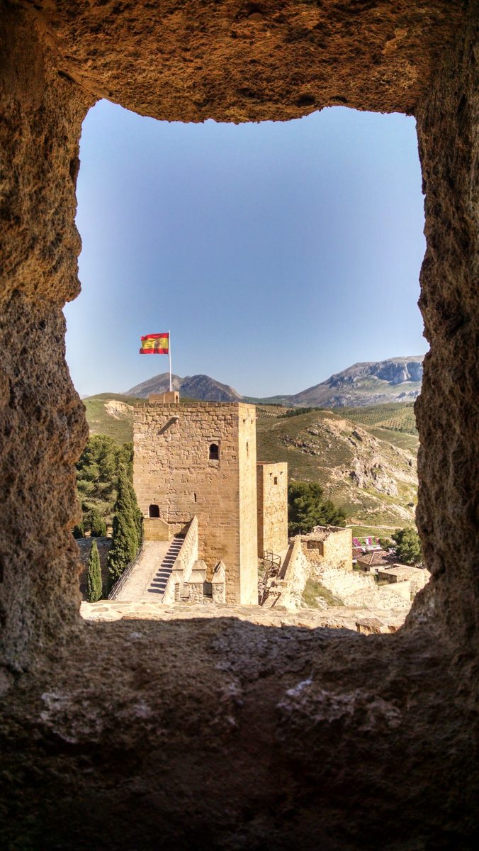 View out rough window of old fortress looking at fortress, Spanish flag, and mountains