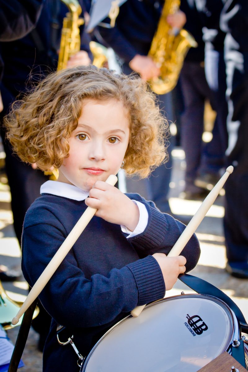 Young girl in band uniform with curly hair and small drum and sticks looks at camera