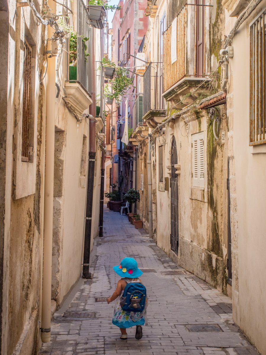 Small girl in blue hat and dress walks down alley with old Italian buildings