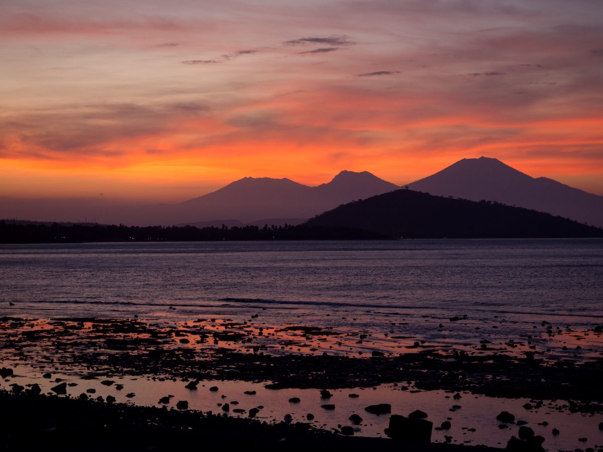 Silhouettes of hill and rocks across water with volcanos in distance at sunset