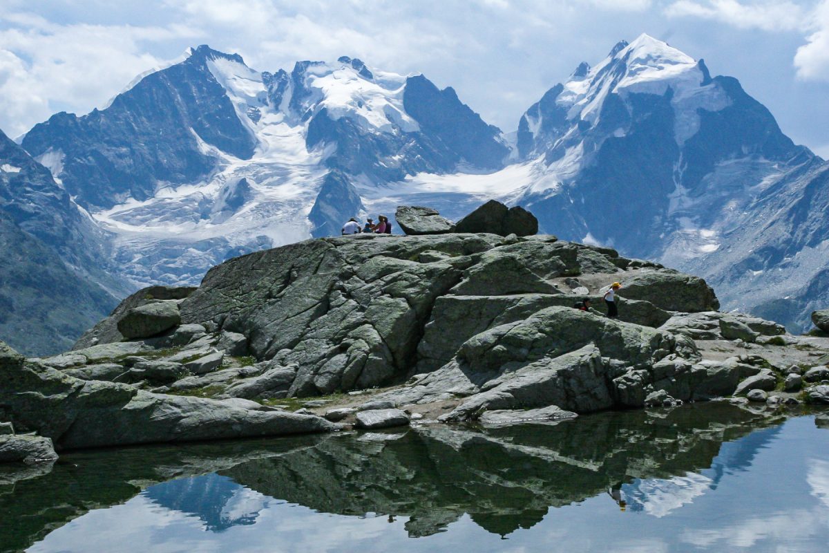 Hikers sit on rock above pond and look out to huge mountains with snow