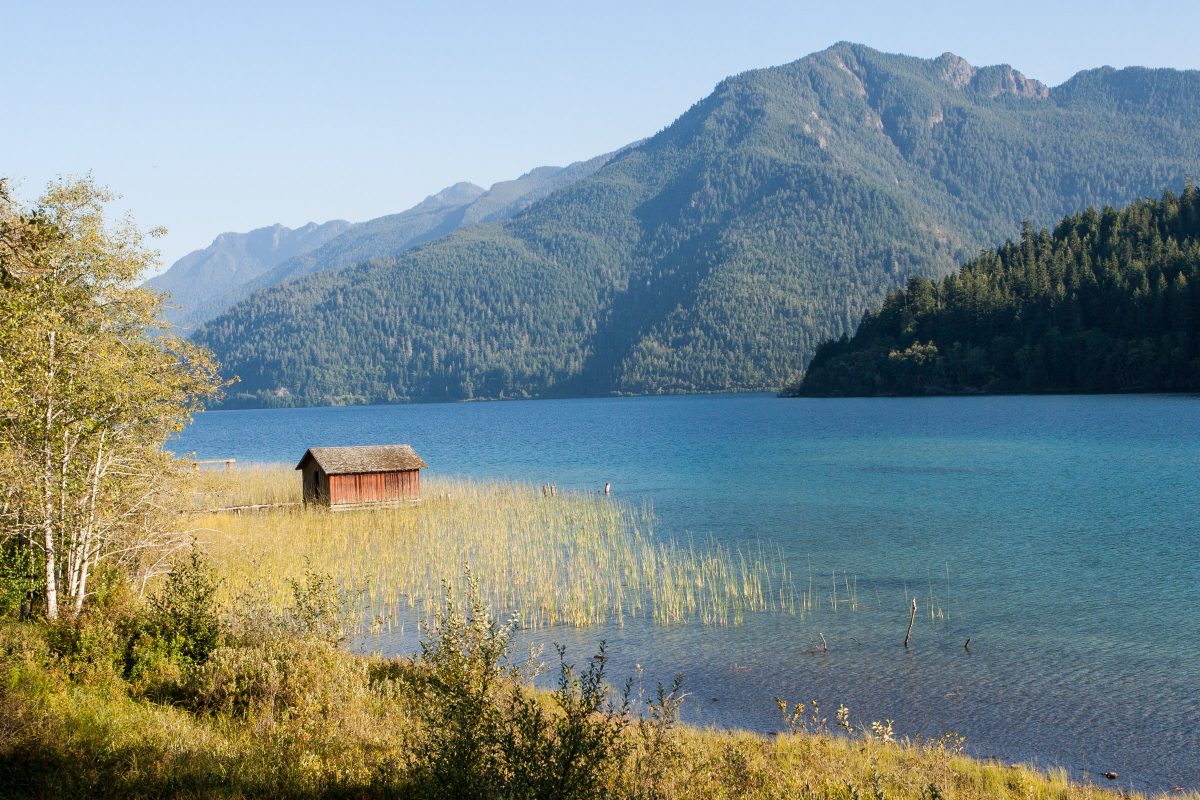 Red boathouse and tan grasses on blue lake with mountains behind