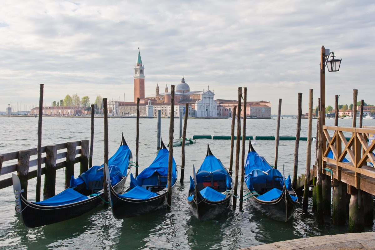 Black gondolas with blue covers tied to poles. View across water to island with church and other buildings