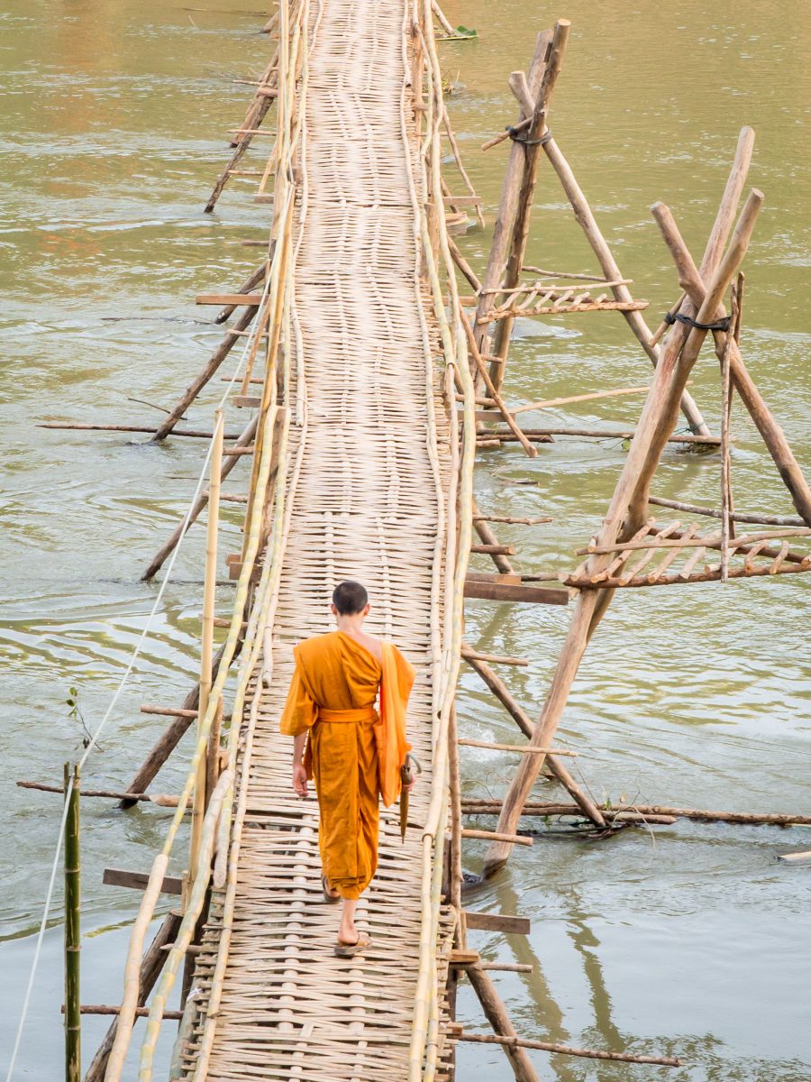 Monk in orange robe crosses long bamboo brige over river