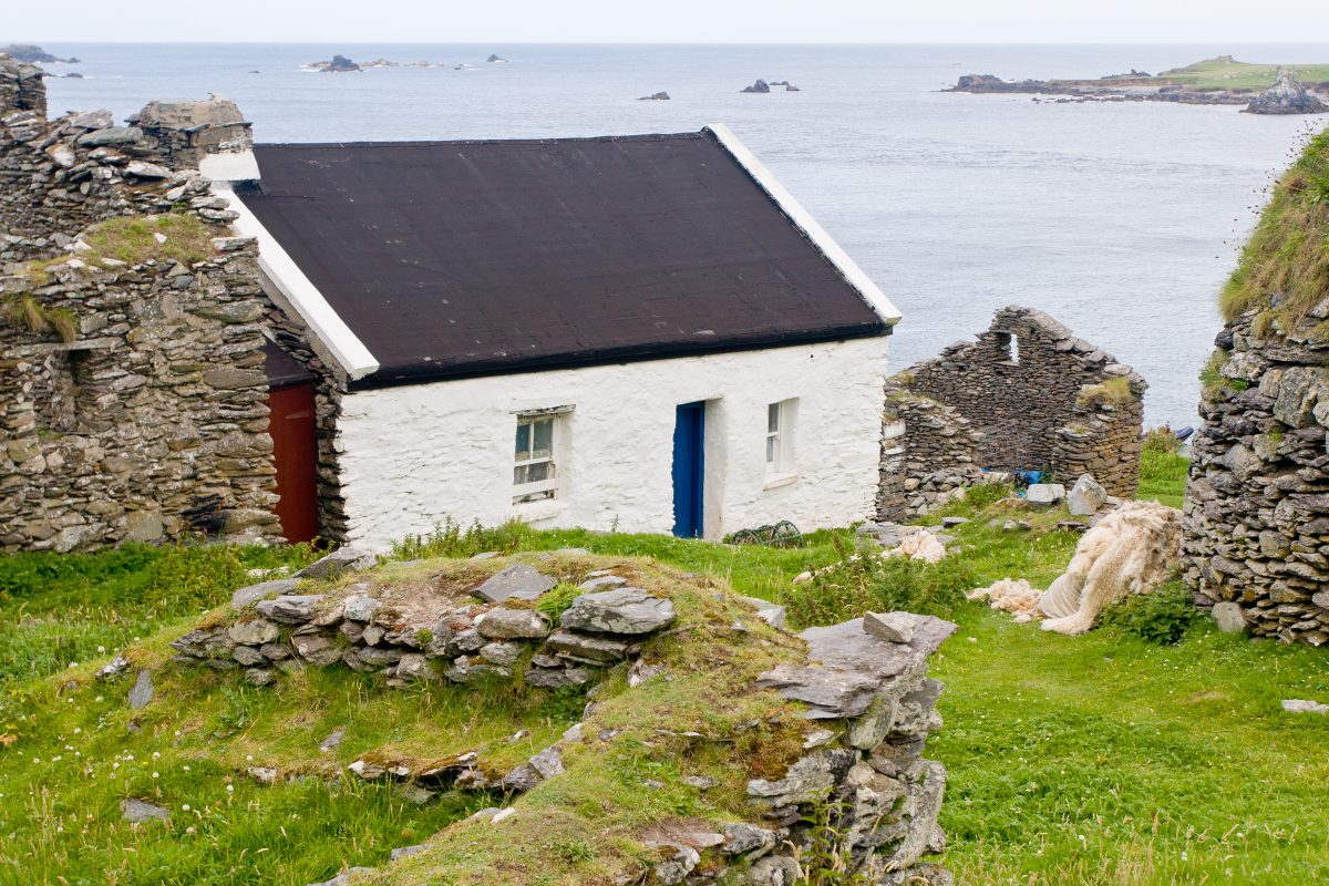 White stone house with black roof amid other ruined stone houses looking out toward ocean
