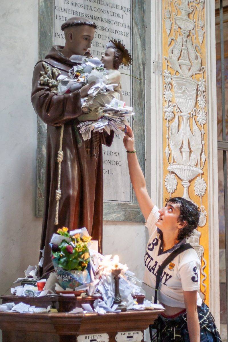 Girl leans up to place note into pile of other notes around statue of saint in brown robe