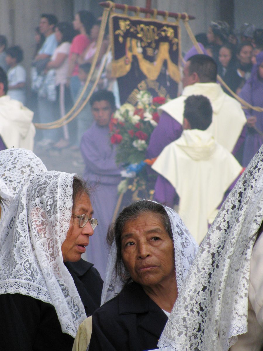 Two Mayan women in lace headscarfs in foreground. Men in purple carrying flowers and banners in background.
