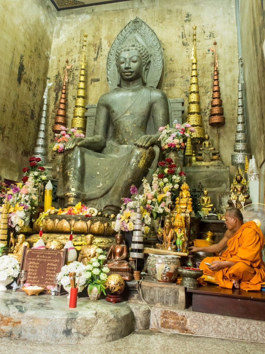 Monk in orange robe sits cross-legged next to large Buddha statue and many small Buddhas and flower decorations