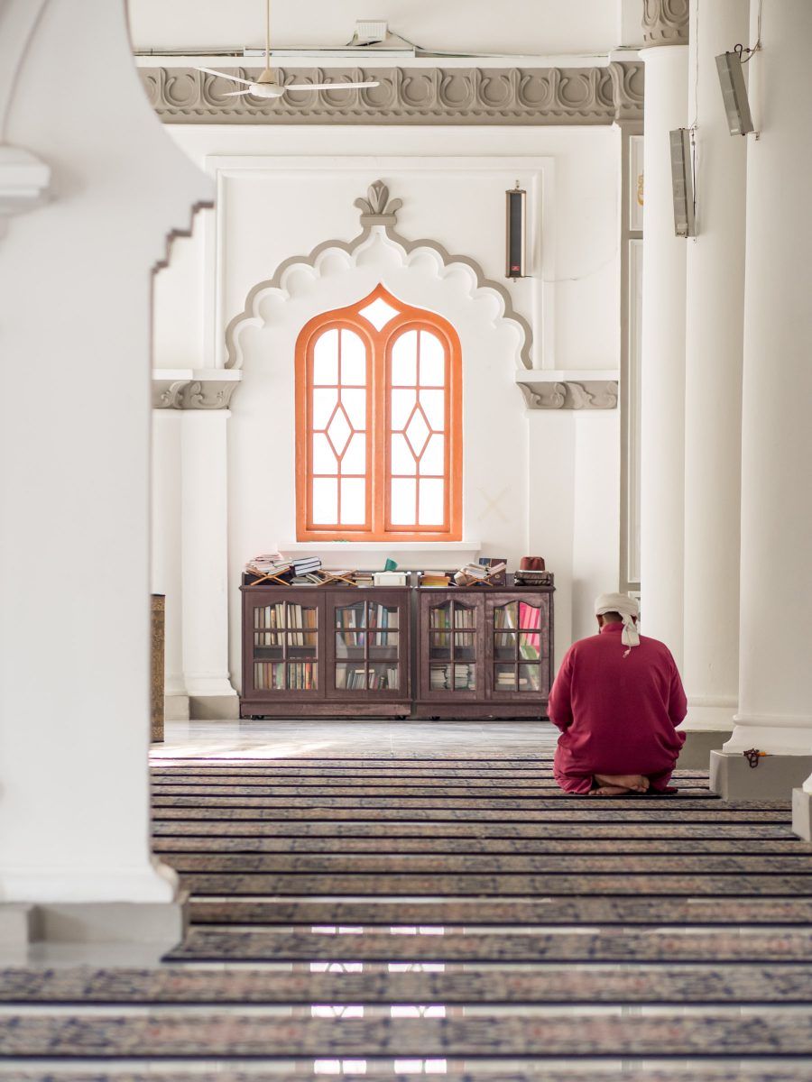 Man in red robe kneels in prayer amid white columns, case of books, and Moorish-style window