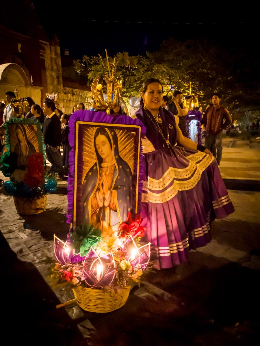 Woman in fancy purple dress poses for photo next to image of Virgin Mary and flowers at night