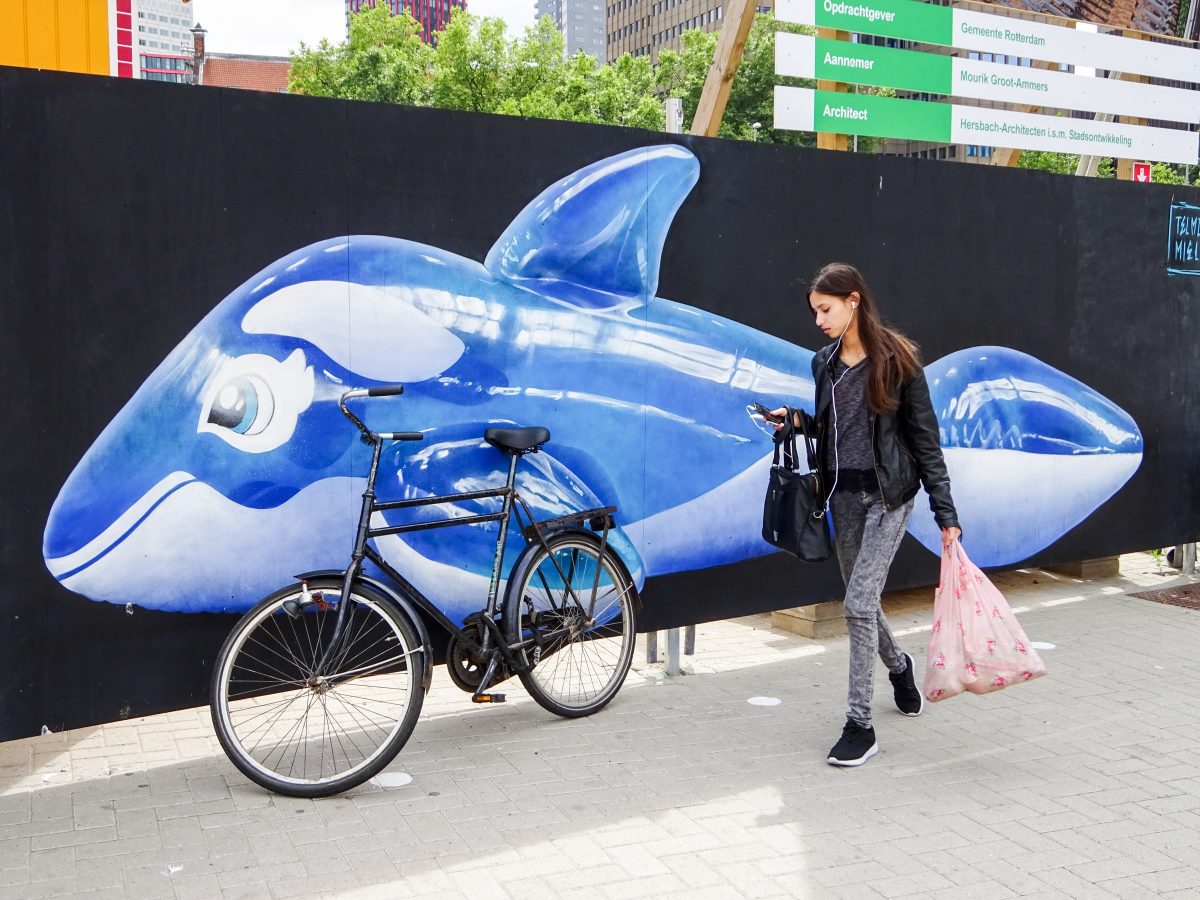 Woman in black carrying bag passes in front of bike and a mural of a blue plastic whale