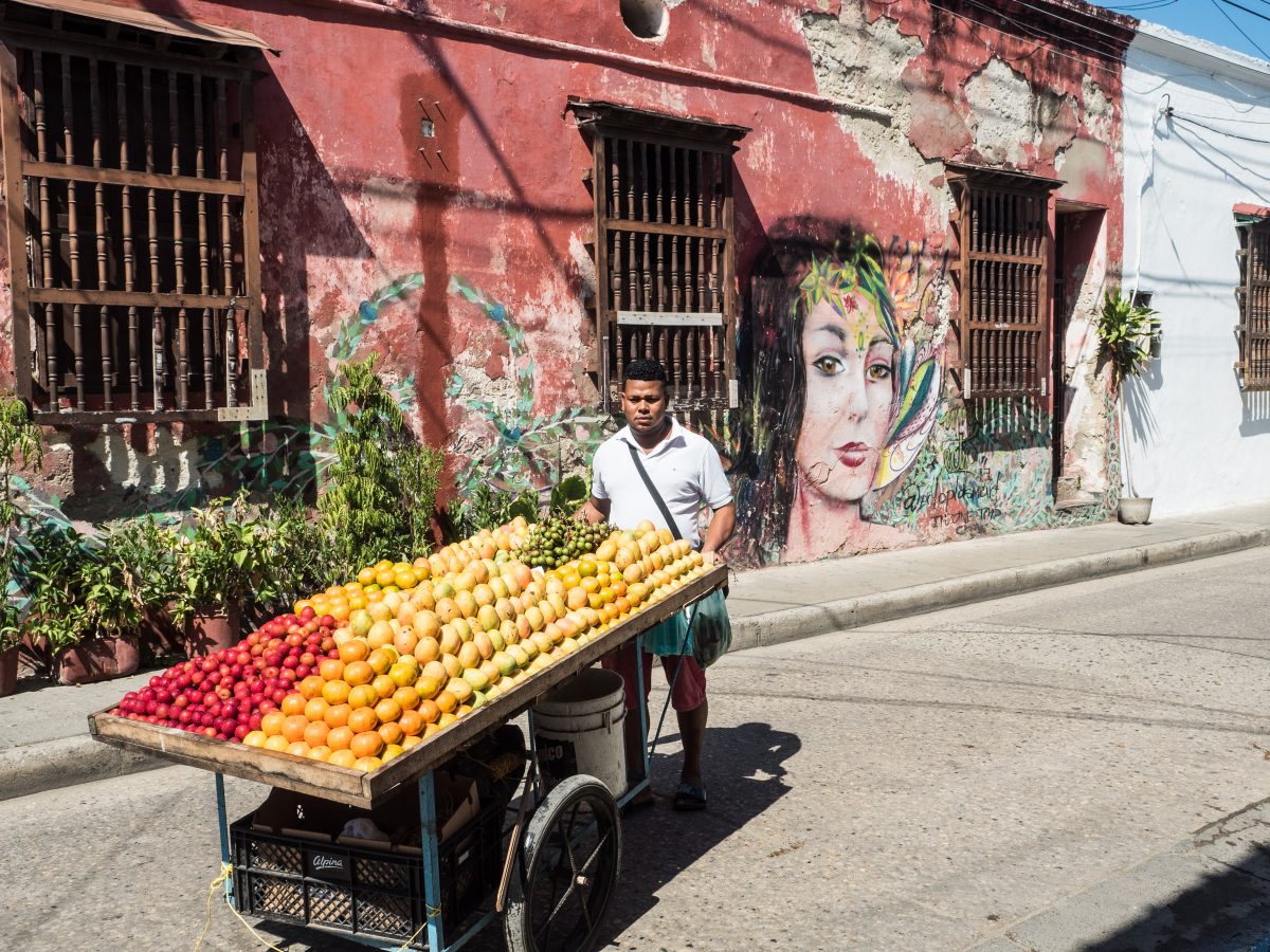 Man pushes cart piled with different fruits down road in front of crumbling red building with mural of a woman's face